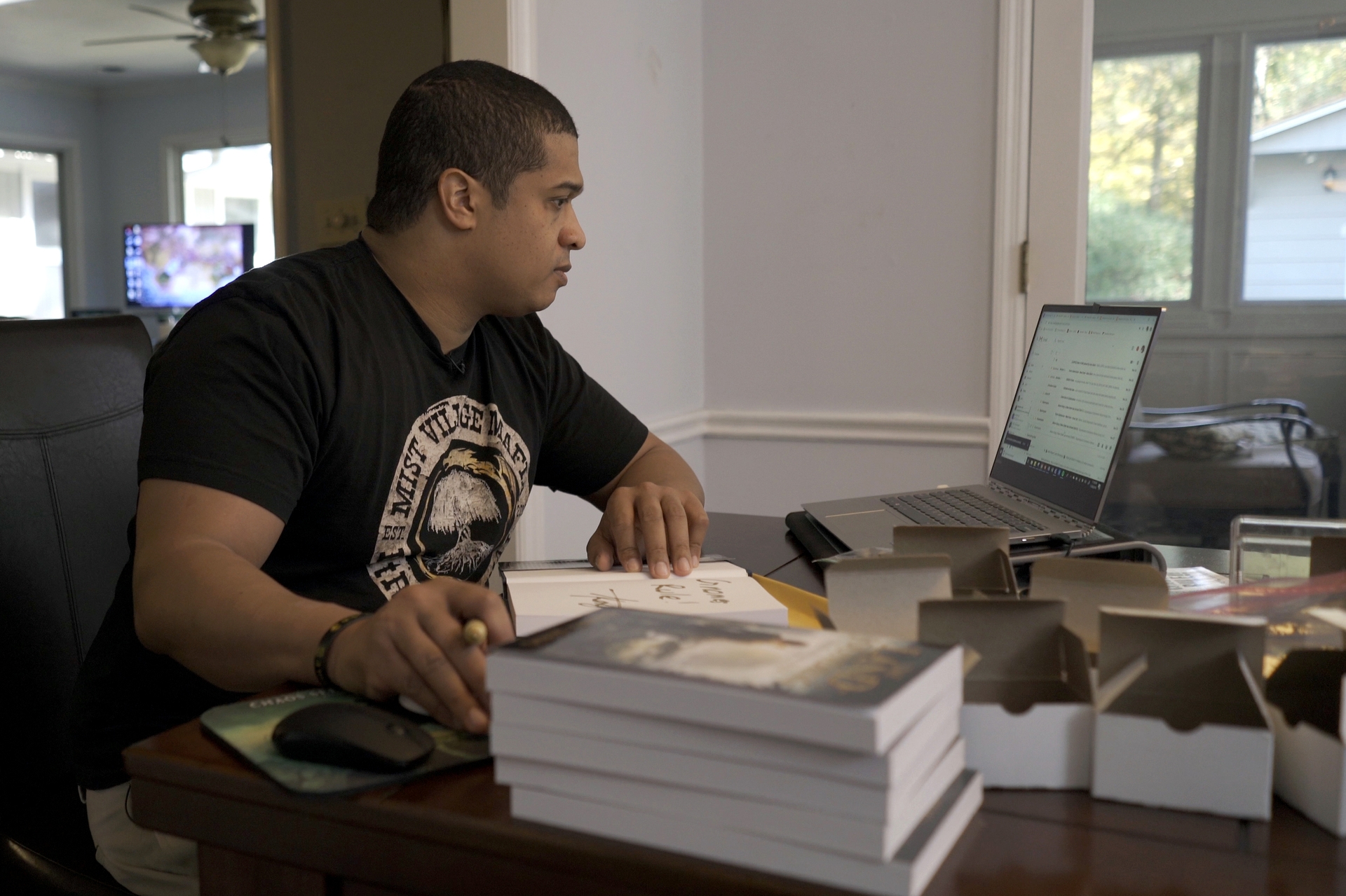 A man sits at a desk in front of a laptop computer. A book is open in front of him.  A stack of books is in the foreground of the image.