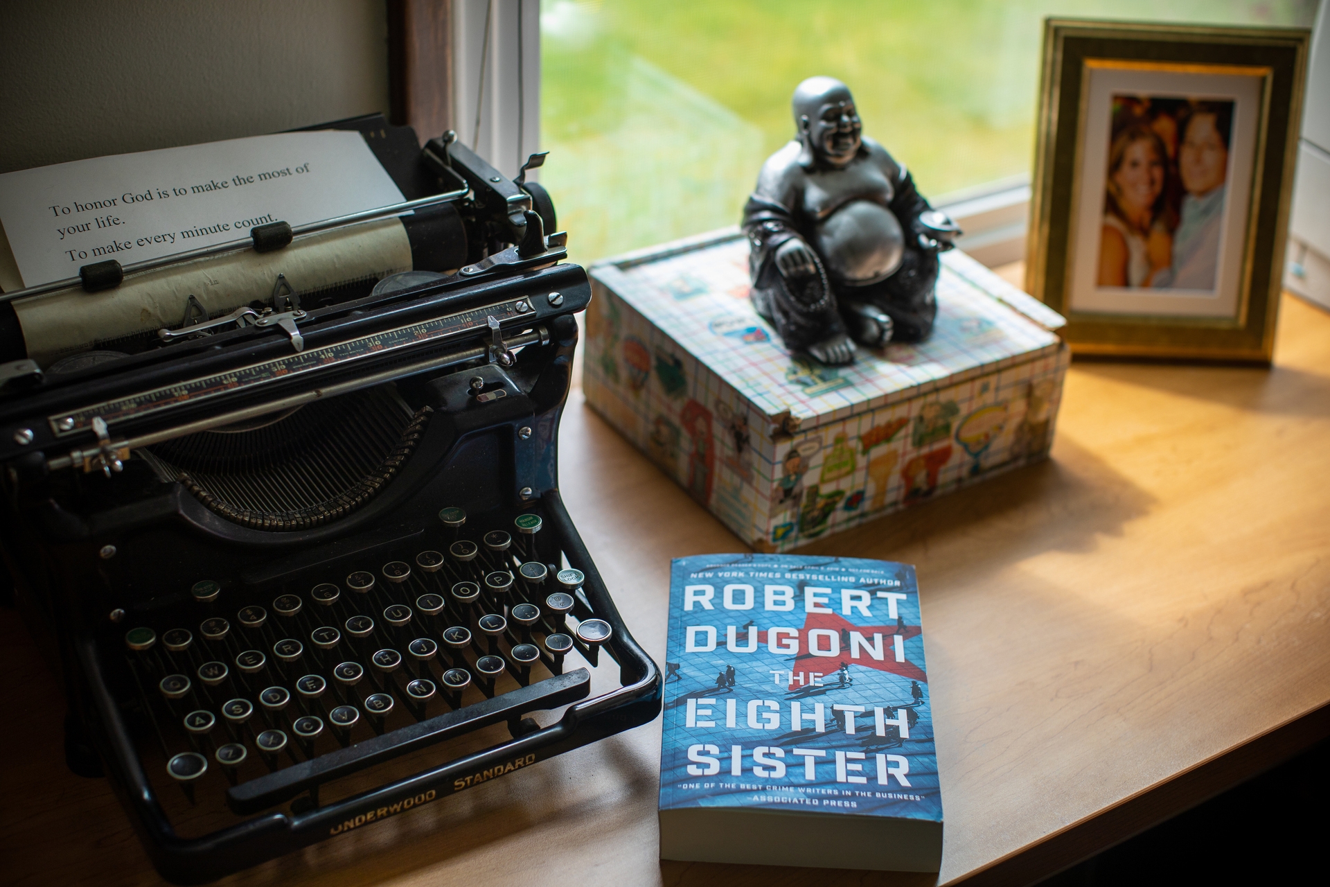 Author Robert Dugoni's workspace within his home office, a refurbished typerwriter sits near a window. To it's left, a small box topped with a Buddha figurine, a copy of his book "The Eighth Sister" and a framed photograph. 