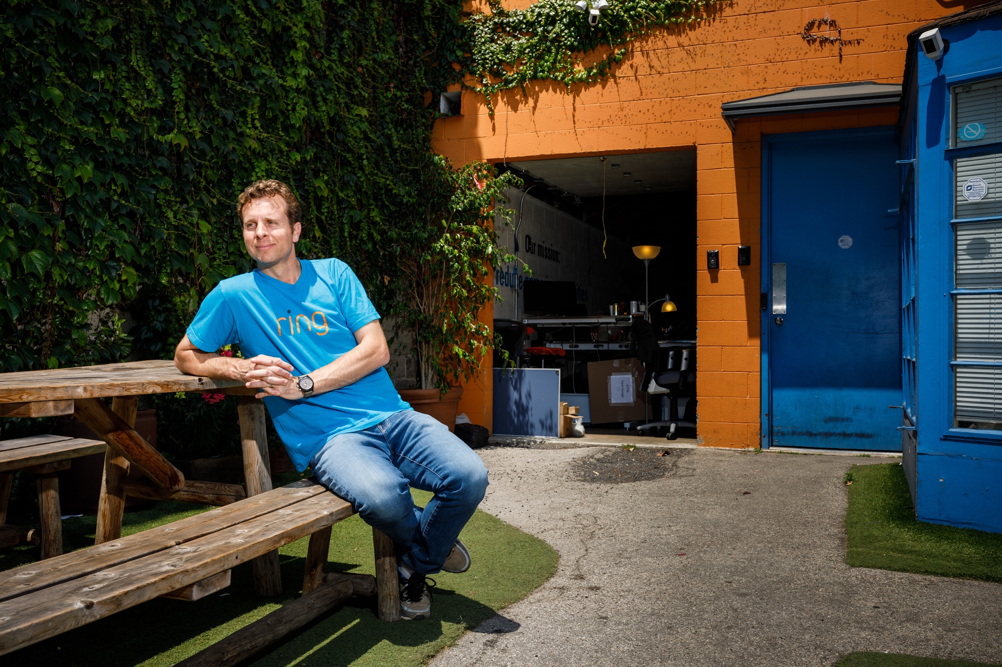 Jamie Siminoff, founder of Ring, sits at a picnic table at Ring headquarters in Santa Monica, CA. He is in a blue t-shirt and jeans, and is looking to the left. 