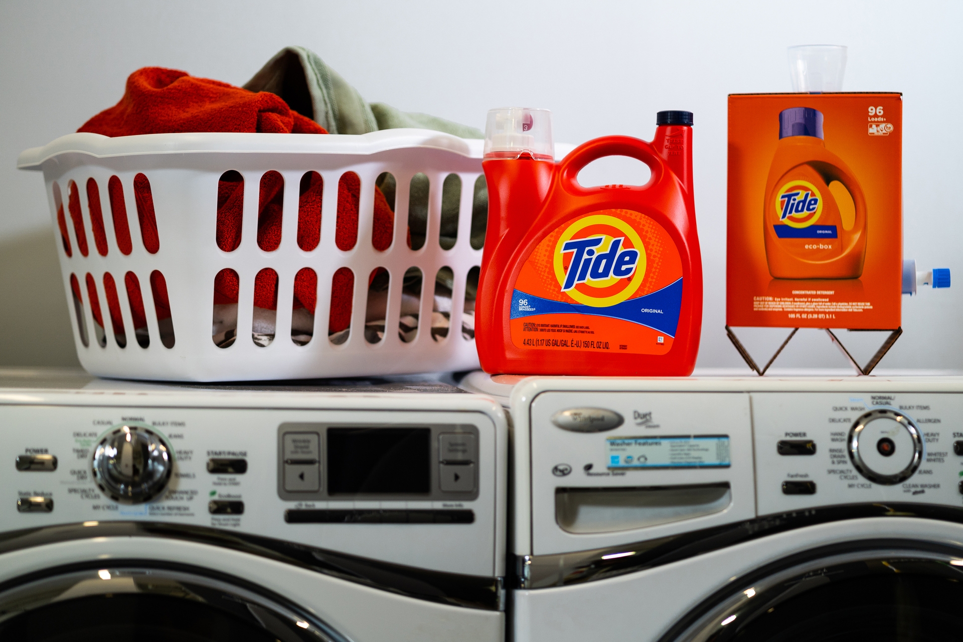 A basket of laundry sits atop a washer and dryer. Next to the laundry basket there are two containers of Tide laundry detergent. One is made of plastic. The other is made of cardboard and sits atop two triangular legs.