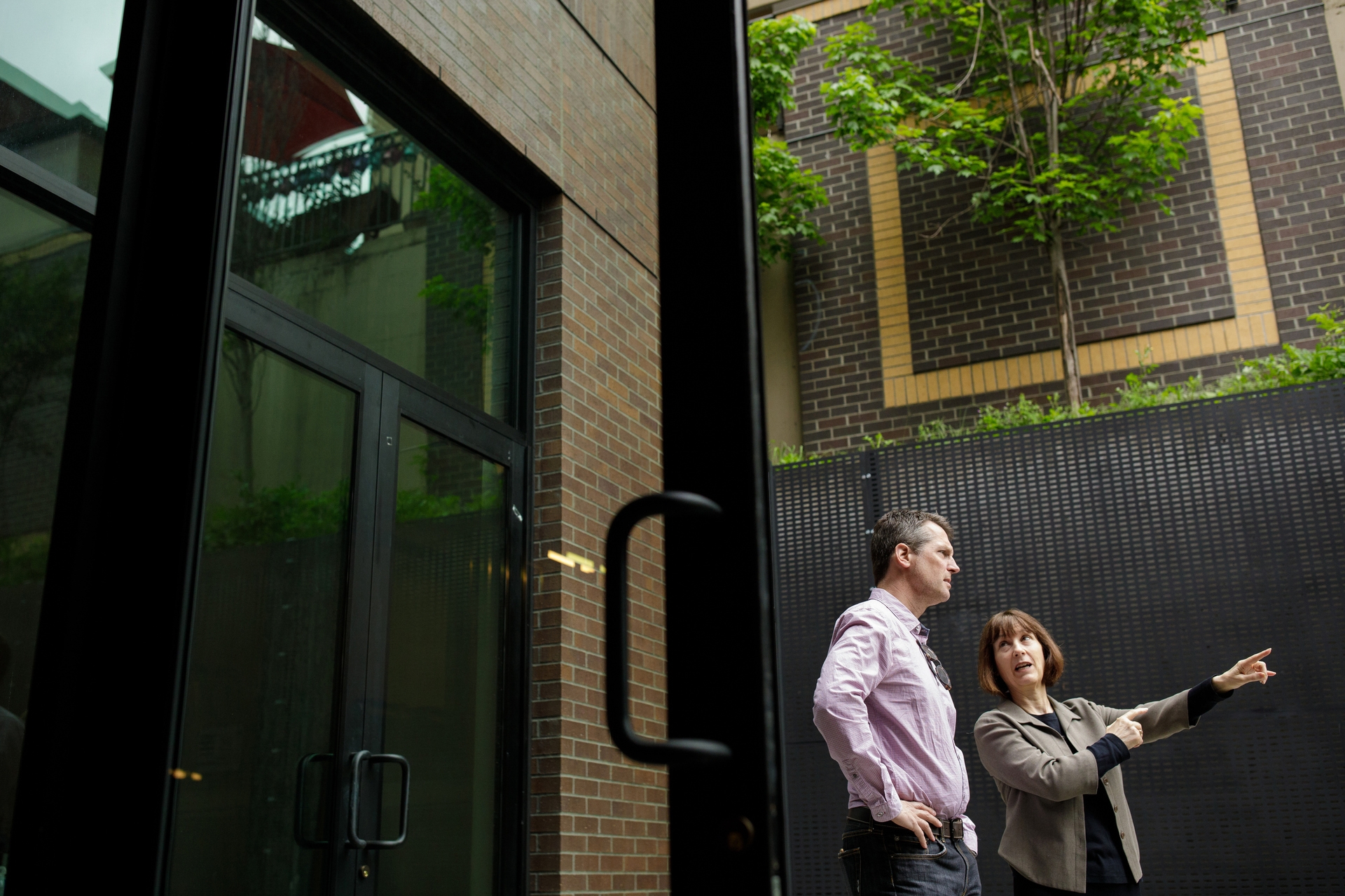 Seen through a glass door, a man and a woman stand in an open-air courtyard. The woman points to something to the right. A tree and a brick wall are at the top of the image.