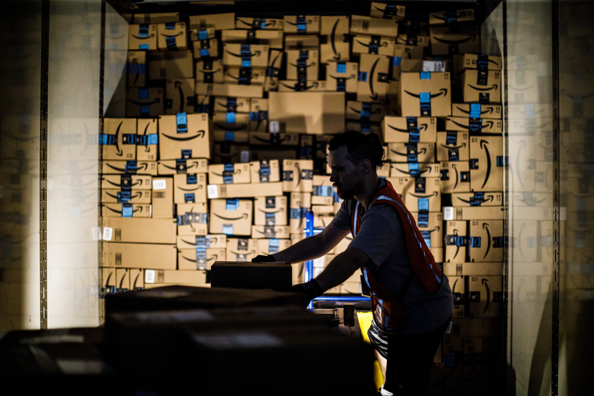 An Amazon associate, dramatically back lit by his work light, stacks shipping boxes labeled with the Amazon smile to the ceiling of a semi trailer.