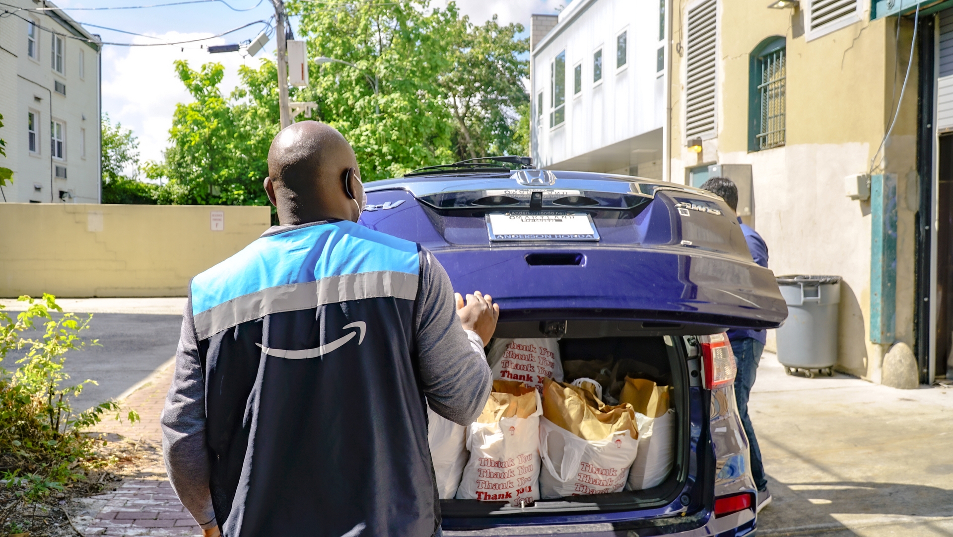 A man in a vest with the Amazon smile logo closes the back of a car filled with bags of groceries.