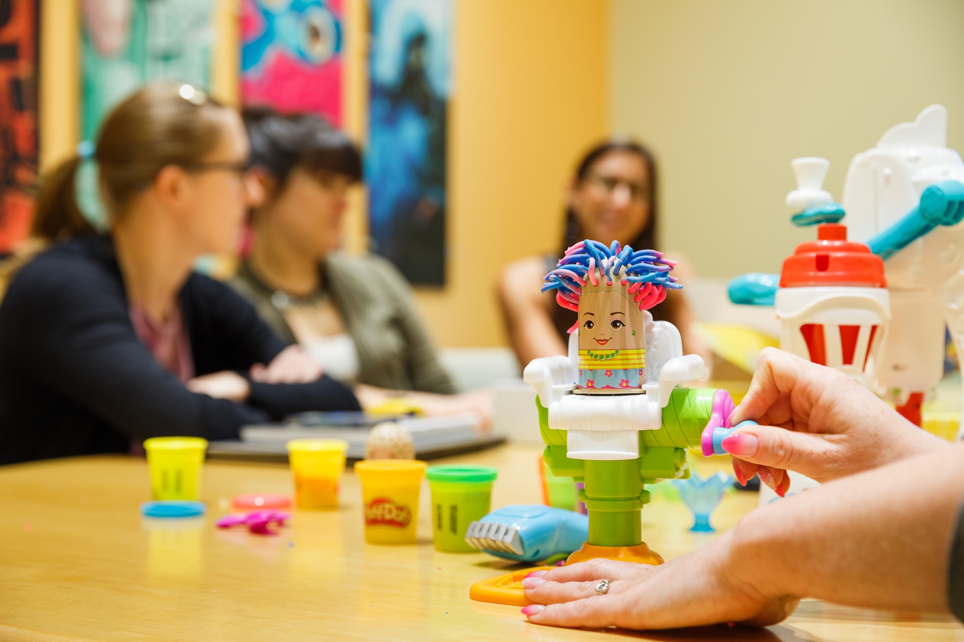 In the foreground, a person turns a crank to demonstrate a Play-Doh toy. In the background, three women sit at a conference table.