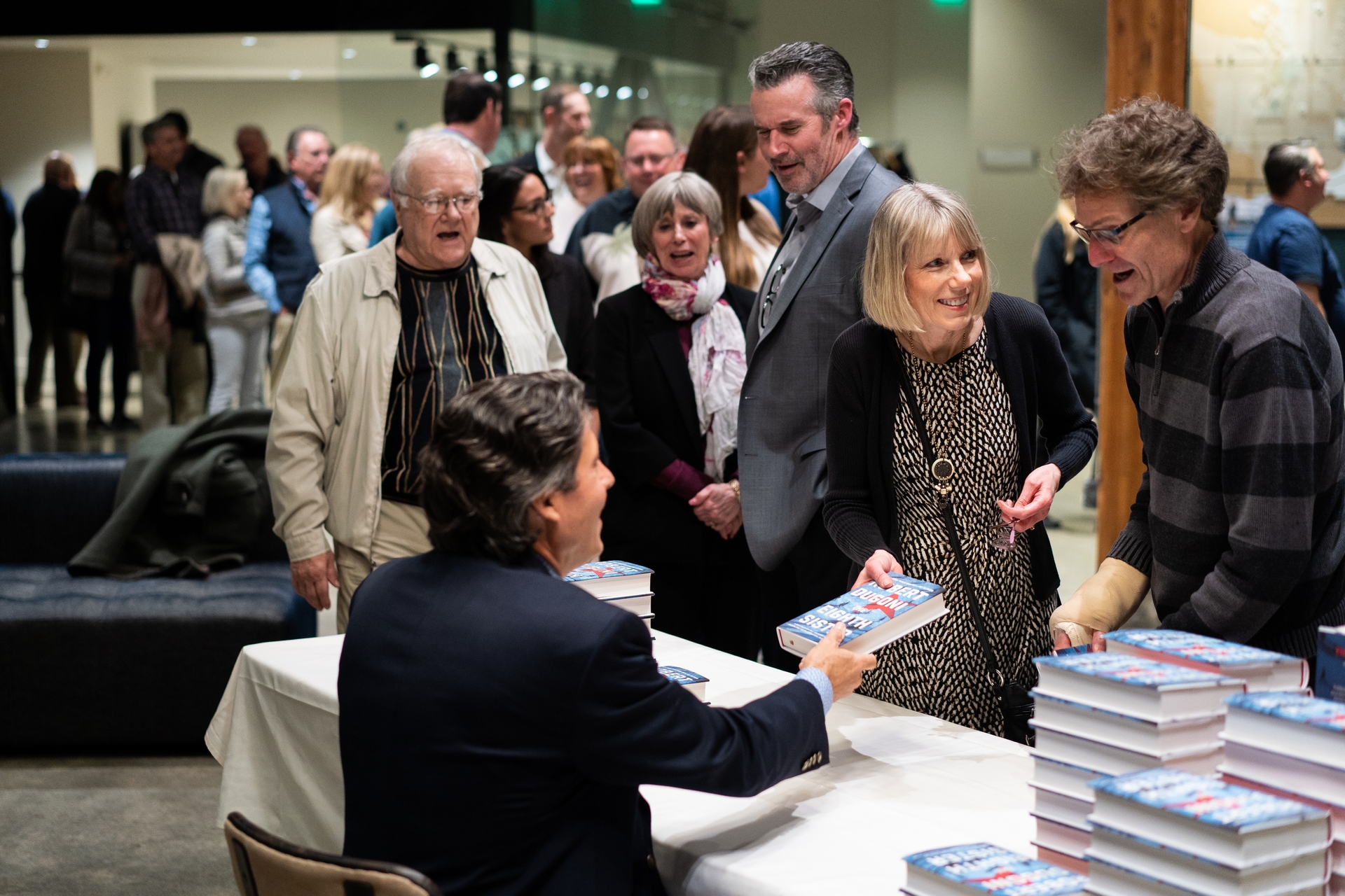 Author Robert Dugoni sits at a table at a book signing event. Fans stand near him, leaning in to get a book signed and share a few words.