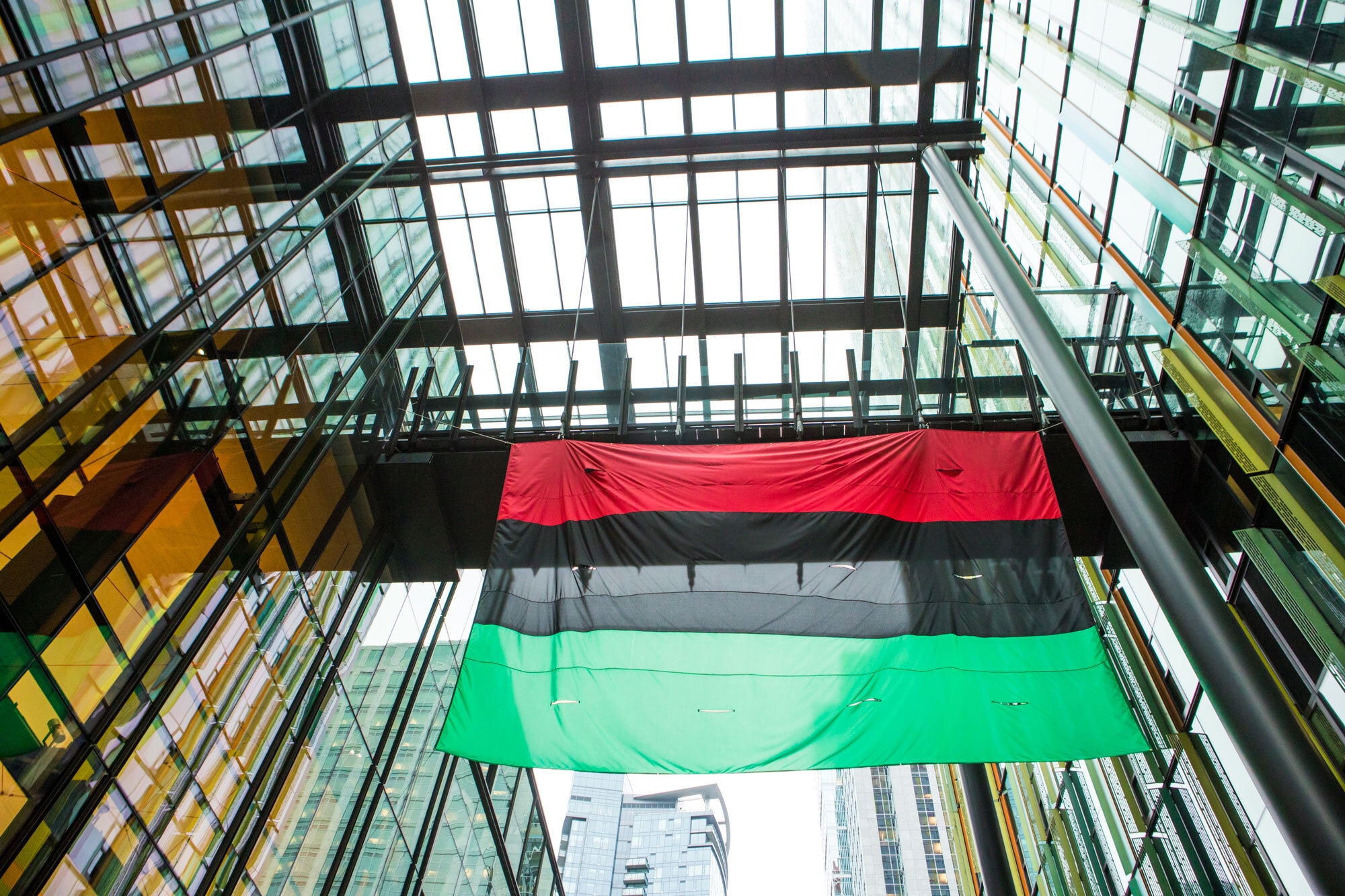 A flag with red, black, and green horizontal stripes flies from a skybridge between two office buildings.