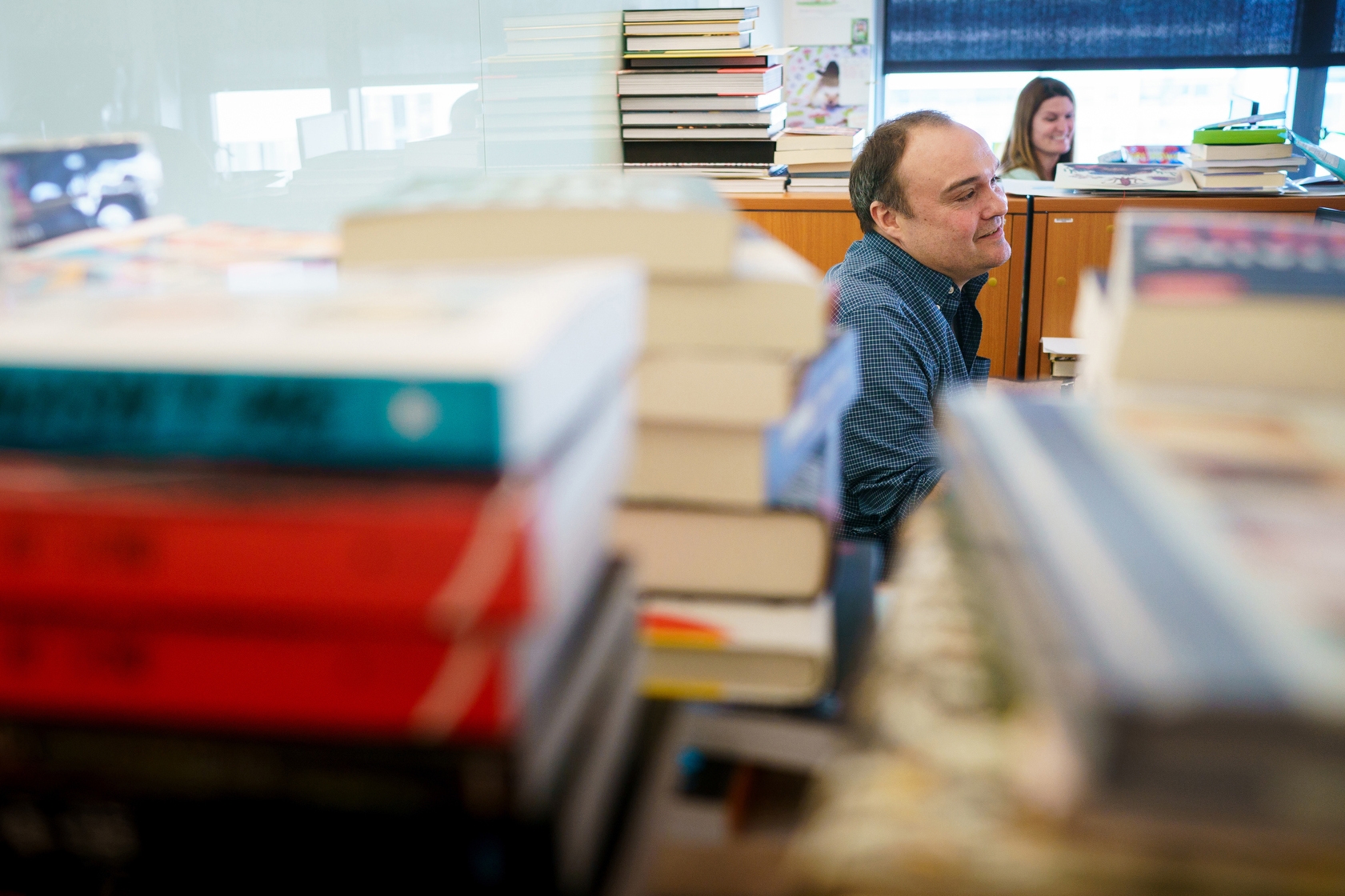 A seated man is mostly obstructed by stacks of books in the foreground of this image. He wears a dark button-down shirt and has brown hair. In the background is a woman with long brown hair as well as more stacked books on top of book shelves.