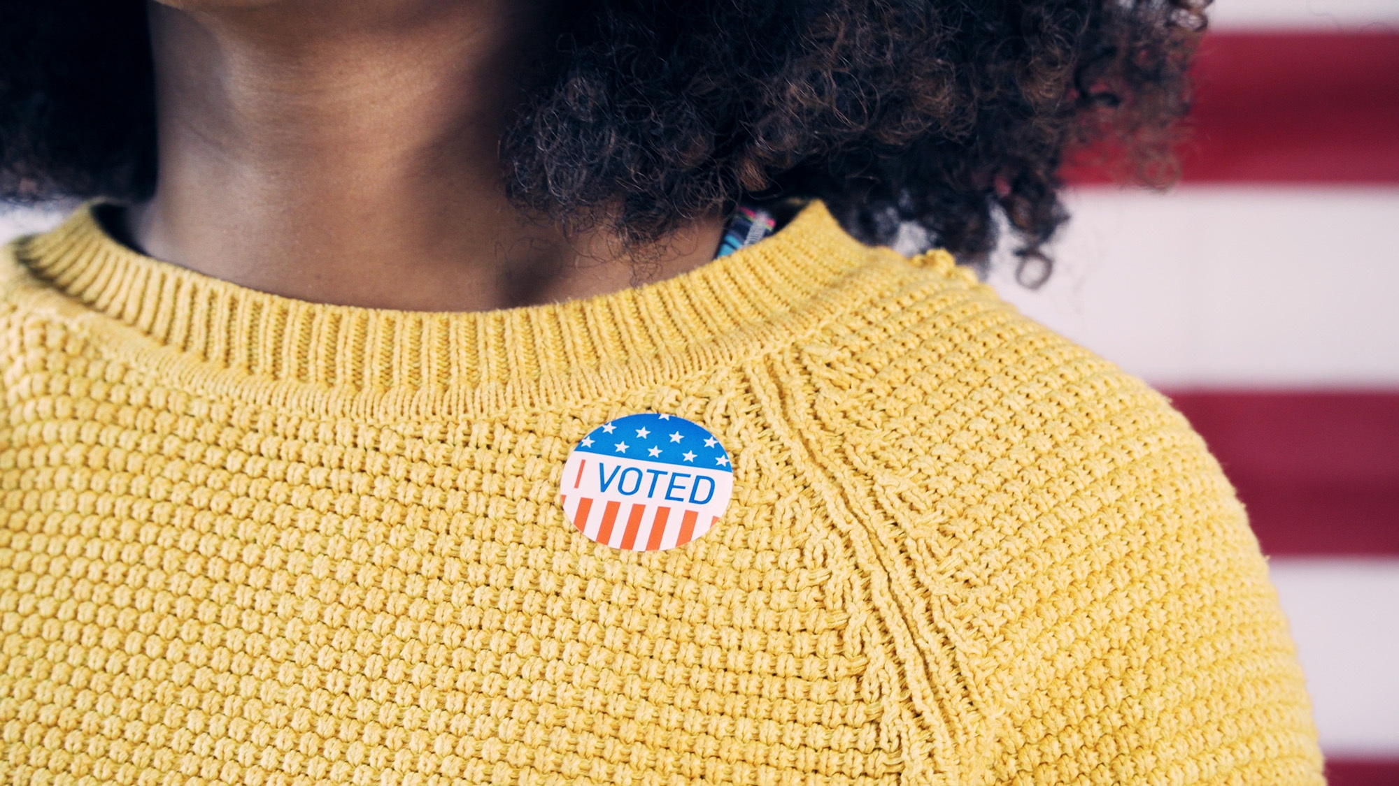 A woman with brown skin and curly hair wears a yellow sweater. Upon that sweater, she wears a sticker that states, "I VOTED" with white stars on blue background, and red and white stripes. 